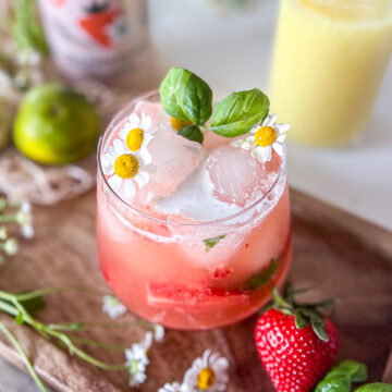 A refreshing strawberry ginger mocktail garnished with basil and chamomile flowers, served on a wooden tray surrounded by chamomile flowers with strawberries, a mason jar full of ginger juice and a lime nearby.