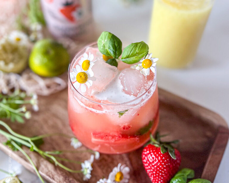 A refreshing strawberry ginger mocktail garnished with basil and chamomile flowers, served on a wooden tray surrounded by chamomile flowers with strawberries, a mason jar full of ginger juice and a lime nearby.
